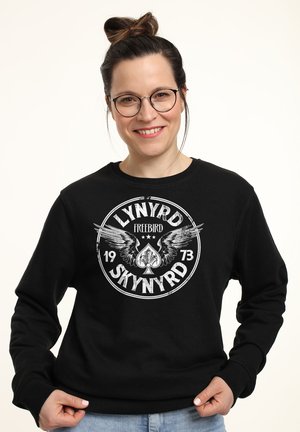 Sweatshirt noir avec un graphisme circulaire portant le texte "LYNYRD SKYNYRD", des ailes et un design de pique. Col rond et coupe décontractée.