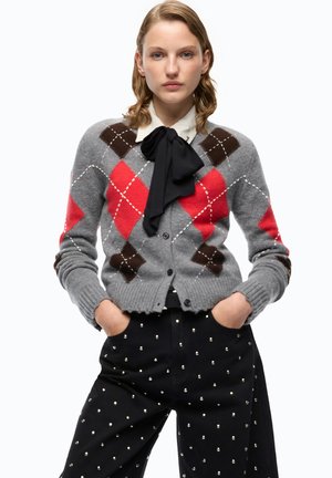 Donna che indossa un cardigan grigio con motivo argyle rosso e marrone, camicia bianca con papillon nero e pantaloni neri a pois bianchi, mani nelle tasche.