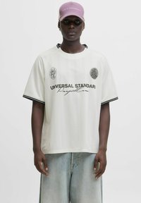 Camiseta de manga corta blanca con detalles negros, que presenta el texto "UNIVERSAL STANDARD" y logotipos. Combinada con unos vaqueros anchos de color azul claro.
