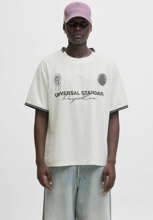 Camiseta de manga corta blanca con detalles negros, que presenta el texto "UNIVERSAL STANDARD" y logotipos. Combinada con unos vaqueros anchos de color azul claro.