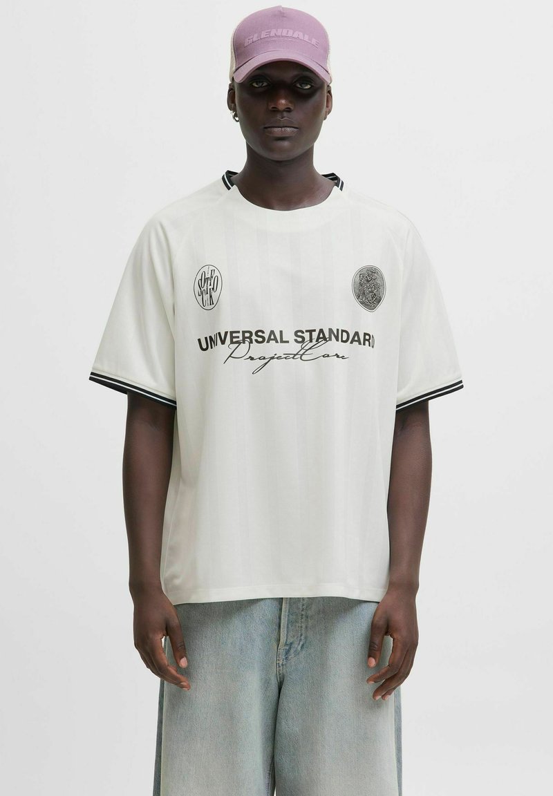 Camiseta de manga corta blanca con detalles negros, que presenta el texto "UNIVERSAL STANDARD" y logotipos. Combinada con unos vaqueros anchos de color azul claro.