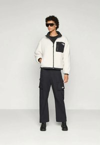 Osoba u bijelom dukserici na zatvaranje s crnom džepom na prsima, crnim kabastim hlačama s više džepova, crnim čizmama i tamnim naočalama, stoji.