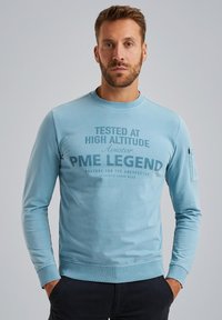 Hellblaues Sweatshirt mit Rundhalsausschnitt, langen Ärmeln und einem Druck auf der Vorderseite. Verfügt über eine Tasche am linken Ärmel und gerippte Bündchen.