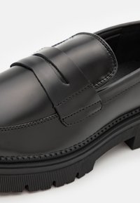 Black leather loafers with a stitch design, featuring a raised seam, a thick ridged sole, and a smooth, glossy finish.