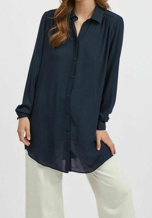 Granatowa, długorękawowa koszula na guziki z kołnierzem, marszczeniem na ramionach i zaokrąglonym dołem, zestawiona z jasnymi spodniami o szerokich nogawkach.