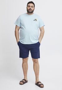 Světle modré bavlněné tričko s grafikou palmy, spárované s námořnickými šortkami. Hnědé sandály dotvářejí neformální outfit.