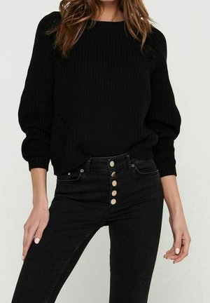 Femme portant un pull en maille noir avec des manches bouffantes et un jean noir taille haute avec cinq boutons visibles à l'avant.