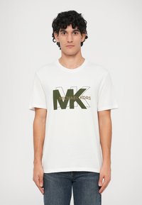 Jauns vīrietis ar cirtainiem melniem matiem, tērpies baltā T-kreklā ar lielu olīvu zaļu "MK" un brūnu "Michael Kors" tekstu, apvienojot to ar zilām džinsām.