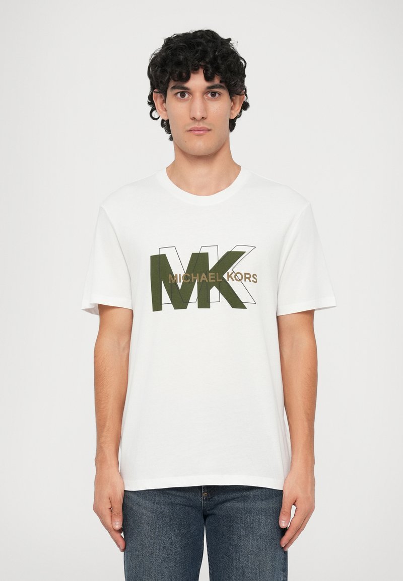 Jauns vīrietis ar cirtainiem melniem matiem, tērpies baltā T-kreklā ar lielu olīvu zaļu "MK" un brūnu "Michael Kors" tekstu, apvienojot to ar zilām džinsām.