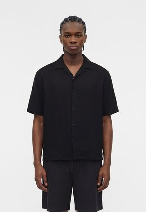 Νεαρός άνδρας με πλεξούδες, που φοράει μαύρο κοντομάνικο πουκάμισο με κουμπιά και μαύρα σορτς, στέκεται μπροστά σε λευκό φόντο.