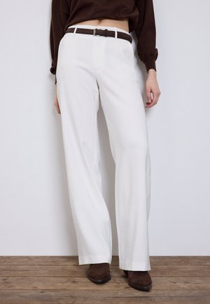 Frau trägt weiß-weit geschnittene, hoch taillierte Hosen, einen dunkelbraunen Gürtel, ein dunkelbraunes Langarmshirt und dunkelbraune Schuhe und steht auf einem Holzboden.