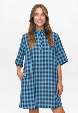 Donna con capelli lunghi che sorride, indossando un vestito a quadri blu e nero, di lunghezza al ginocchio, con maniche corte e colletto, con una mano in tasca.