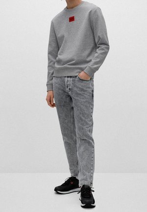 Sudadera gris con un parche de logo rojo en el pecho, combinada con jeans negros deslavados y zapatillas negras con acentos rojos. Estilo simple y casual.