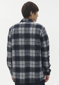 Moški nosi modro, črno in sivo karirano srajco z dolgimi rokavi, obrnjeno proč, z kratkimi rjavimi lasmi in temnimi hlačami.