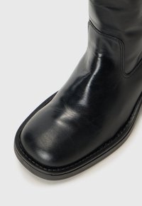 Primer plano de una bota de cuero negro, con un acabado brillante y una punta cuadrada, sobre un fondo blanco liso.