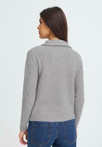 Grå ribbestrikket trøje med krave, lange ærmer og tætsiddende kant, båret over jeans. Blød tekstur med et subtilt mønster.