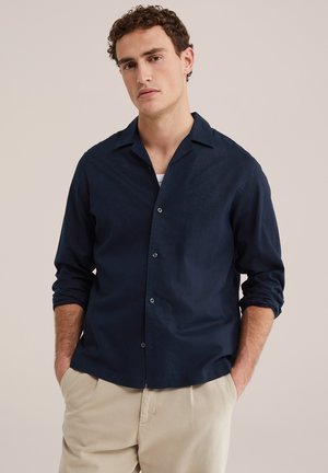 Tânăr cu păr creț, purtând o cămașă bleumarin cu nasturi și pantaloni bej, mâinile în buzunare, fundal neutru.