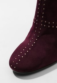 Gros plan sur l'avant d'une botte en daim violet foncé ornée de petits clous métalliques ronds disposés en lignes verticales et diagonales.