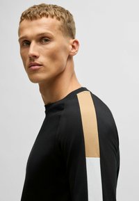Chemise noire à manches longues avec des rayures beige et blanches sur les épaules, dotée d'un col rond et d'une texture lisse. Design minimaliste.