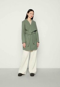 Pikkade mustade juustega naine, kes kannab vööga oliivrohelist mantlit, laiu kreemjaid pükse ja musti kingi, seisab ühevärvilise heleda tausta ees.