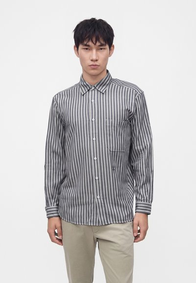 Homme portant une chemise à rayures verticales grises et blanches boutonnée avec un pantalon beige, se tenant devant un fond blanc uni.