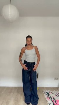 Γυναίκα με πλεγμένα μαλλιά, φοράει γκρι αμάνικη μπλούζα και φαρδιά ναυτικά μπλε παντελόνια, στέκεται σε ένα δωμάτιο με ξύλινο πάτωμα και λευκούς τοίχους.