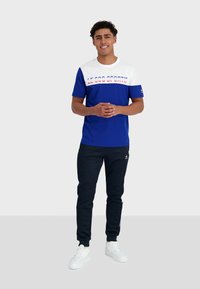 T-shirt bleu et blanc avec le texte "LE COQ SPORTIF", associé à un pantalon noir et des baskets blanches, présentant un logo sur le pantalon et la manche.
