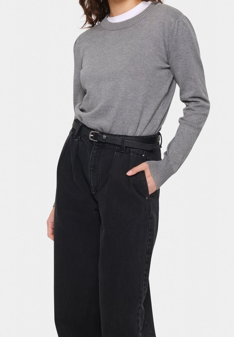Femme portant un pull gris à manches longues rentré dans un jean noir ample taille haute avec une ceinture noire, une main dans la poche.