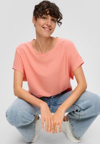 Kurzärmelige Bluse mit Rundhalsausschnitt aus zartem Korallstoff. Kombiniert mit hochgeschnallten, hellen, weit geschnittenen Jeans und weißen Sneakers.