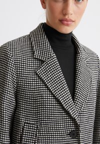 Blazer cu un model houndstooth alb-negru, cu rever în formă de notch, închidere cu un singur buton și țesătură texturată, purtat peste un pulover negru cu guler înalt.