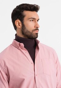 Lichtroze button-up shirt, gedragen over een donkerbruine geribbelde turtleneck. Het shirt heeft een borstzak en zichtbare stiksels.