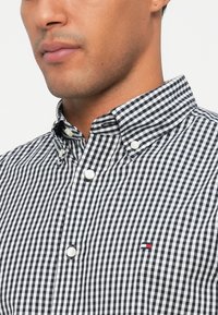 Černobílá kostkovaná košile s límcem na knoflíček, vybavená bílými knoflíky a malým barevným logem na prsou. Bavlněný materiál.