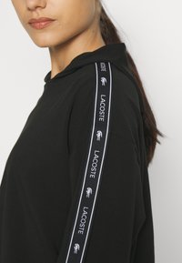 Melna kokvilnas kapuche ar brīvu piegriezumu. Aprīkota ar sasienamu kapuci un kontrastējošām sānu lentēm ar "LACOSTE" un logotipa detaļām.