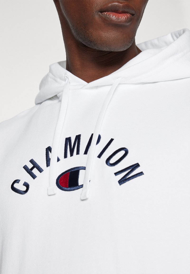 Weißer Kapuzenpullover aus weichem Material mit navyblauem gesticktem Text "CHAMPION" und einem runden Logo in Rot und Blau. Zugbänder an der Kapuze.