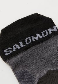 Calcetín negro y gris hecho de un tejido suave, con una sección acolchada y un logo "SALOMON" en gris claro cerca de la parte superior.