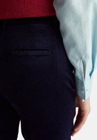 Navy corduroy pants with a back pocket, paired with a light blue textured shirt featuring buttoned cuffs. Red knit sweater visible above.