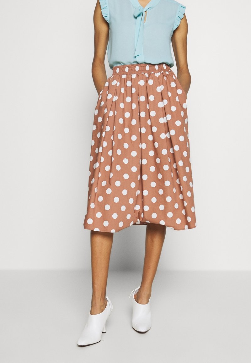 Femme portant un chemisier sans manches bleu et une jupe marron à pois blancs jusqu'aux genoux, assortis à des chaussures à talons blanches.