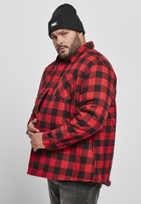 Rød og svart rutete flanellskjorte med to frontlommer, knapplukking og lange ermer, kombinert med en svart lue.