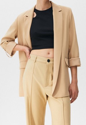 Personne portant un blazer beige et un pantalon avec manches retroussées et un top court noir, main reposant sur la hanche sur fond uni.