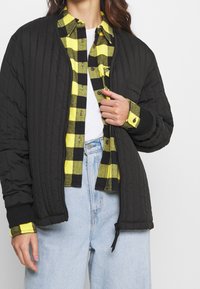 Giacca nera trapuntata con polsini a coste, indossata sopra una camicia a scacchi gialla e nera, con una maglietta bianca sotto e jeans di denim chiaro.