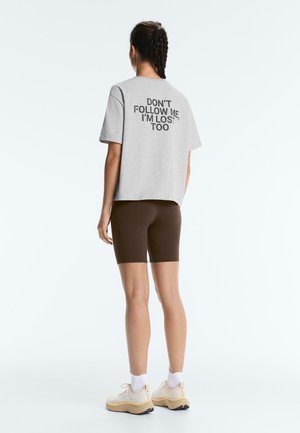 Osoba s vrkočmi má na sebe sivé tričko s nápisom „NE following ME, I'm LOST TOO“, hnedé šortky, biele ponožky a béžové tenisky, otočená chrbtom.