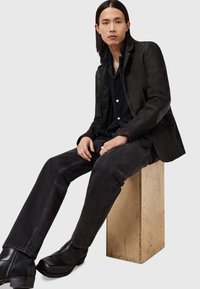 Blazer de cuero negro con una textura suave, combinado con una camisa negra de botones y jeans de mezclilla oscura. El modelo se sienta sobre un bloque de madera.