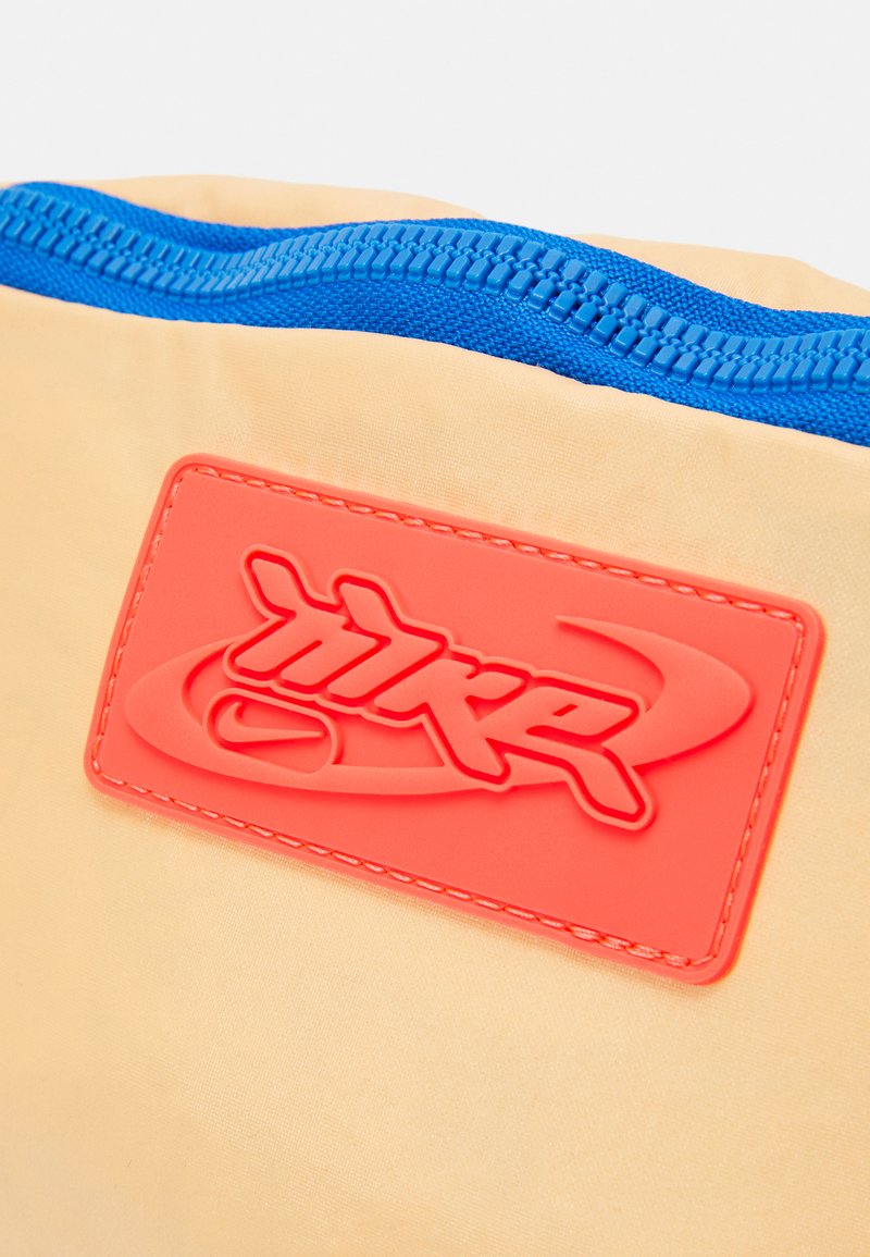 Pochette en tissu jaune avec une fermeture éclair bleue et un patch de logo texturé en orange vif. Texture lisse, forme rectangulaire, avec des détails de couture.