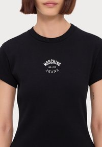 Svart ribbad t-shirt med korta ärmar och rund halsringning, med vit broderad text "MO5CHINO 86-23 JEANS" på framsidan.