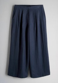 Pantaloni bleumarin cu picior larg, pliuri, talie elastică și buzunare laterale, atârnați pe un fundal deschis, simplu.