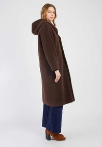 Brązowy, kapturowy płaszcz wykonany z teksturowanej tkaniny, charakteryzujący się prostym krojem i bocznymi kieszeniami. Połączony z ciemnoniebieskimi szerokimi spodniami i brązowymi obcasami.