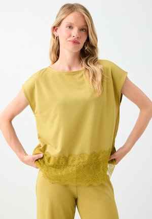 Blond vrouw met een gele losse top met kanten detail aan de zoom en bijpassende broek, staand met de handen in de zij tegen een witte achtergrond.