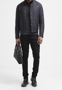 Czarna pikowana kurtka na suwak z teksturowanymi akcentami, noszona na ciemnym swetrze i zestawiona z czarnymi dżinsami oraz błyszczącymi eleganckimi butami. Szara torba trzymana w ręku.