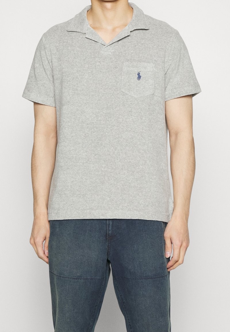 Homme portant un polo gris clair à manches courtes avec un petit logo sombre sur la poche poitrine et un pantalon bleu foncé.