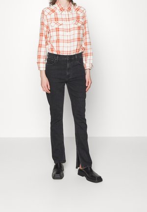 Kvinde iført hvid og orange ternet skjorte med knapper, sorte høj taljede jeans med slidser foran på benene og sorte lædersko uden hæl.